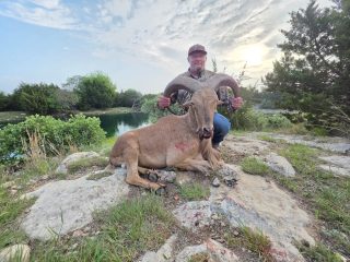 A hunter and an aoudad taken at BarBell Ranch in Sweetwater, TX, beside a tranquil water source.