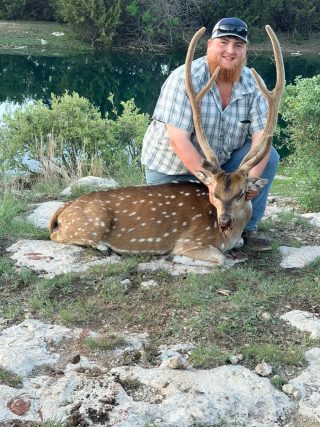 Hunter with a magnificent Axis buck taken at BarBell Ranch in Sweetwater, TX, beside a clear pond.