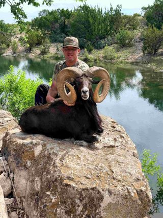 Hunter Chance Tidwell kneels beside his record Midnight Urial, perched on a rock overlooking a pond.