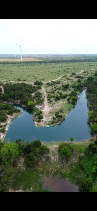 Aerial view: a tranquil clear lake on rolling land with winding roads and wind turbines on the distant horizon.