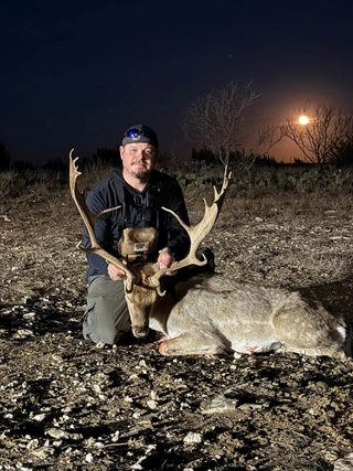 A hunter kneeling with a Fallow buck taken at BarBell Ranch in Sweetwater, TX, illuminated by moonlight.
