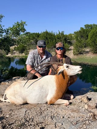 Two hunters with a scimitar oryx taken at BarBell Ranch in Sweetwater, TX, beside a clear pond.